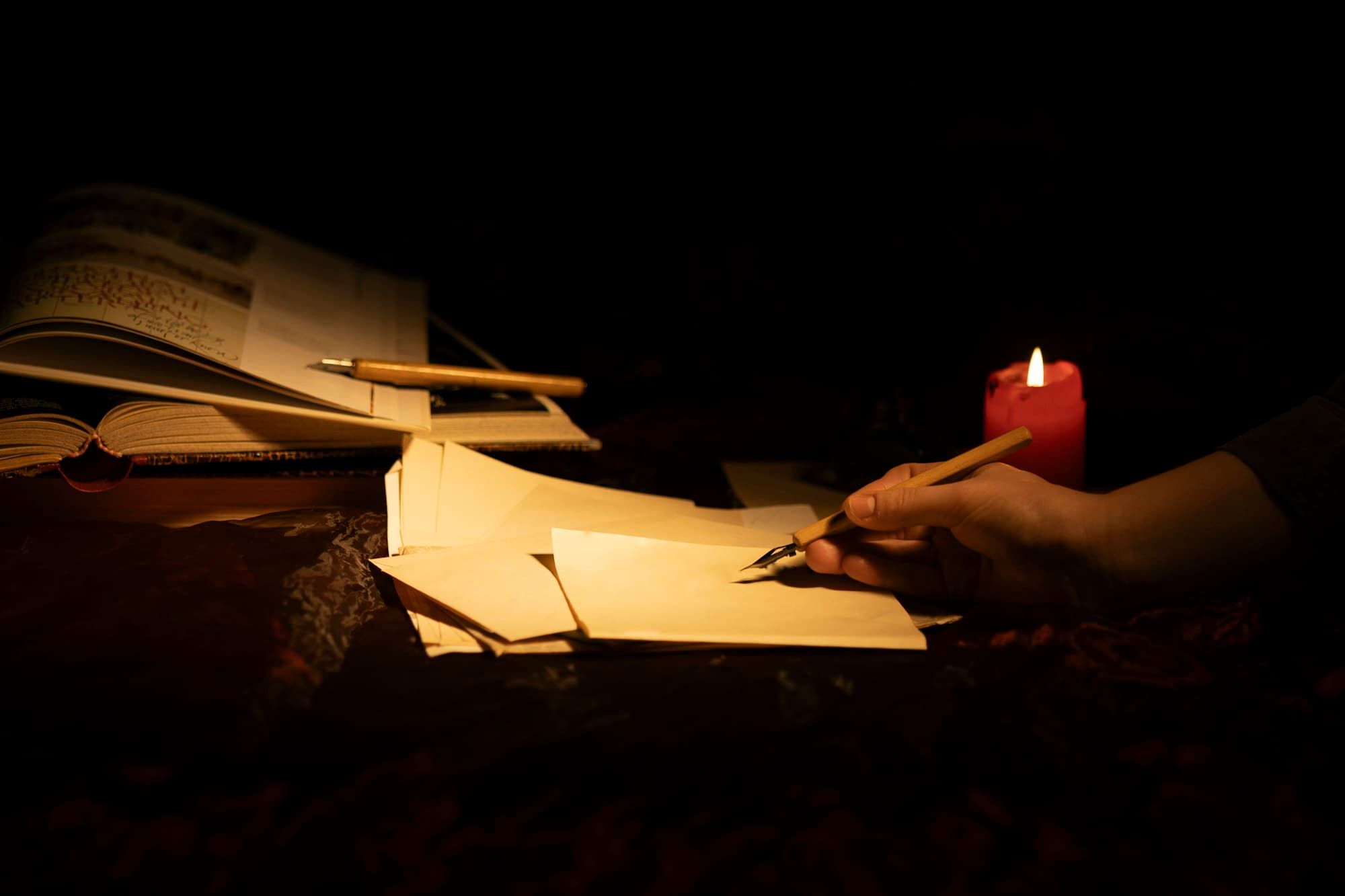 A person writing a letter on paper beside a lit candle.